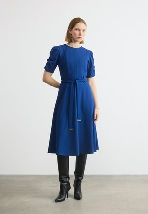 Žena stojaca s rukou za chrbtom, oblečená v modrých šatách po kolená s nafúknutými rukávmi, opaskom v páse a čiernymi čižmami na podpätku.