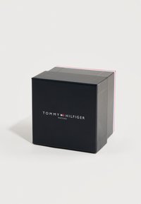 Μαύρο ορθογώνιο κουτί με το λογότυπο Tommy Hilfiger και την επιγραφή "Ρολόγια", τοποθετημένο σε λευκό φόντο.