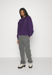 Purple sweatshirt with a crew neck, ribbed hem, and an embroidered logo on the chest, paired with gray cargo pants and beige shoes.