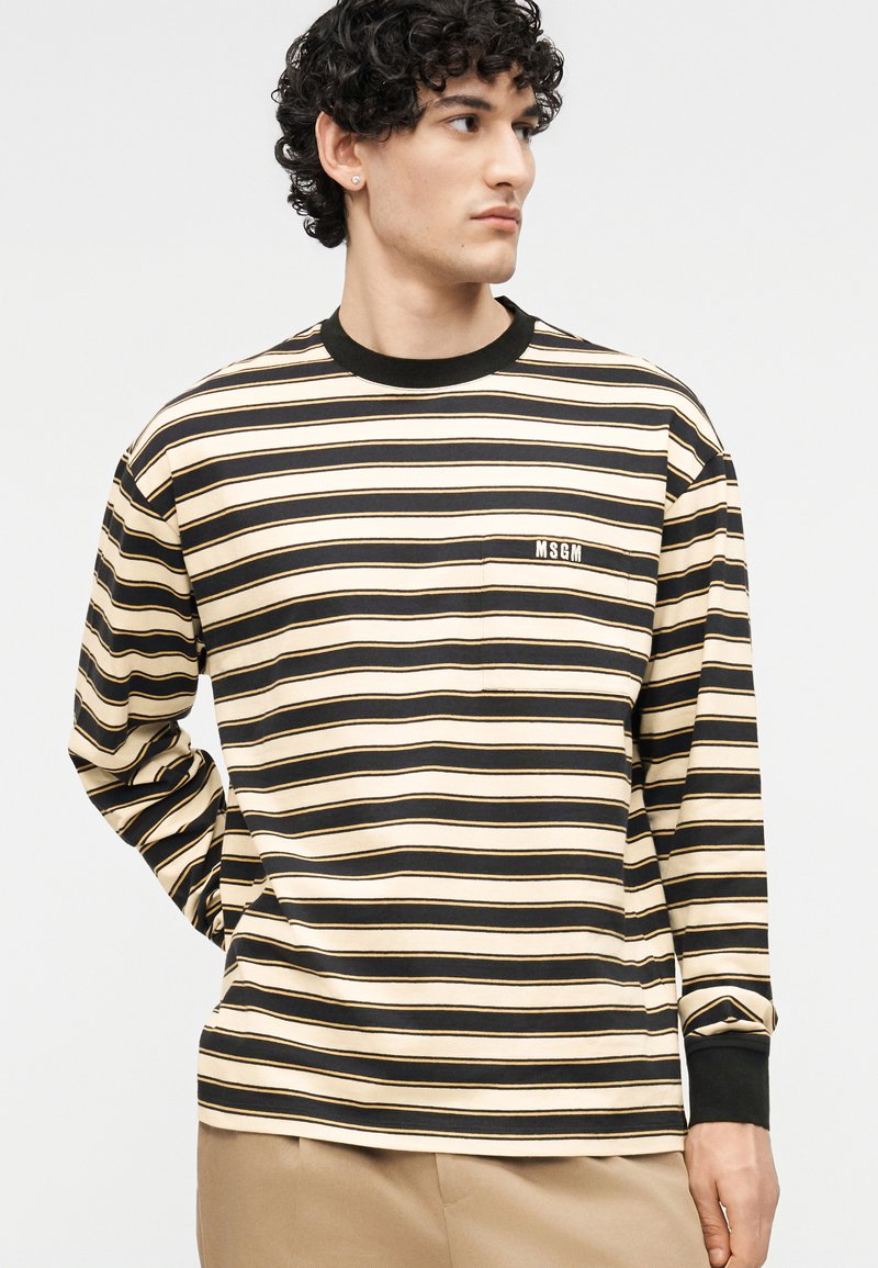 Man die een beige en zwart gestreept shirt met lange mouwen draagt met het MSGM-logo op het borstzakje, kijkt opzij met de hand achter de rug.