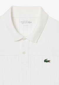 Vit polo skjorta i texturerad tyg med klassisk krage, knapplist och grön broderad krokodil-logotyp på bröstet.