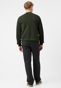 Mężczyzna stoi tyłem, ubrany w ciemnozielony sweter, czarne spodnie i beżowe trampki, na tle jednolitego białego tła.