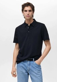 Čierne polo tričko s golierom, krátkymi rukávmi a zapínaním na tri gombíky; spárované so svetlomodrými džínami. Hladká textúra.