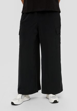 Schwarzfarbene Cargo-Jeans mit weitem Bein und Seitentaschen. Glatte Textur und schlichtes Design. Getragen mit weißen Sneakers.