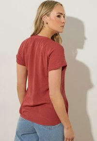 Blusa de manga corta color óxido con un patrón ligeramente texturizado. Cuello redondeado y mangas arremangadas, combinada con jeans de color azul claro.