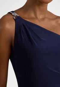 Granatowa sukienka na jedno ramię z drapowanym materiałem, ozdobionym paskiem z kryształkami i gładką fakturą.