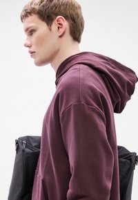 Felpa con cappuccio color marrone scuro con tasca frontale e cappuccio con cordoncino, indossata da una persona che porta uno zaino nero. Tessuto liscio con una vestibilità comoda.