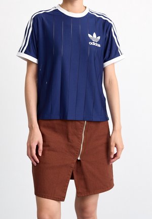 Personen bærer en blå Adidas T-shirt med hvide striber og logo, kombineret med et brunt skørt med lynlås foran og slids.