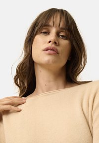 Beiger gerippter Pullover mit weitem Ausschnitt und langen Ärmeln. Der Stoff wirkt weich und texturiert und weist dezente Nähdetails auf.