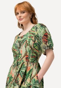 Kurzärmliges Kleid aus grünem Stoff mit einem lebhaften botanischen Druck und roten Akzenten, Knopfdetails und seitlichen Taschen.