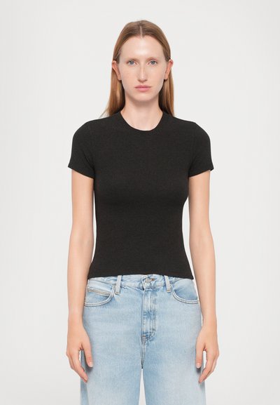 Jeune femme avec des cheveux raides roux, portant un haut noir ajusté à manches courtes et un jean taille haute bleu clair, se tenant devant un fond blanc.