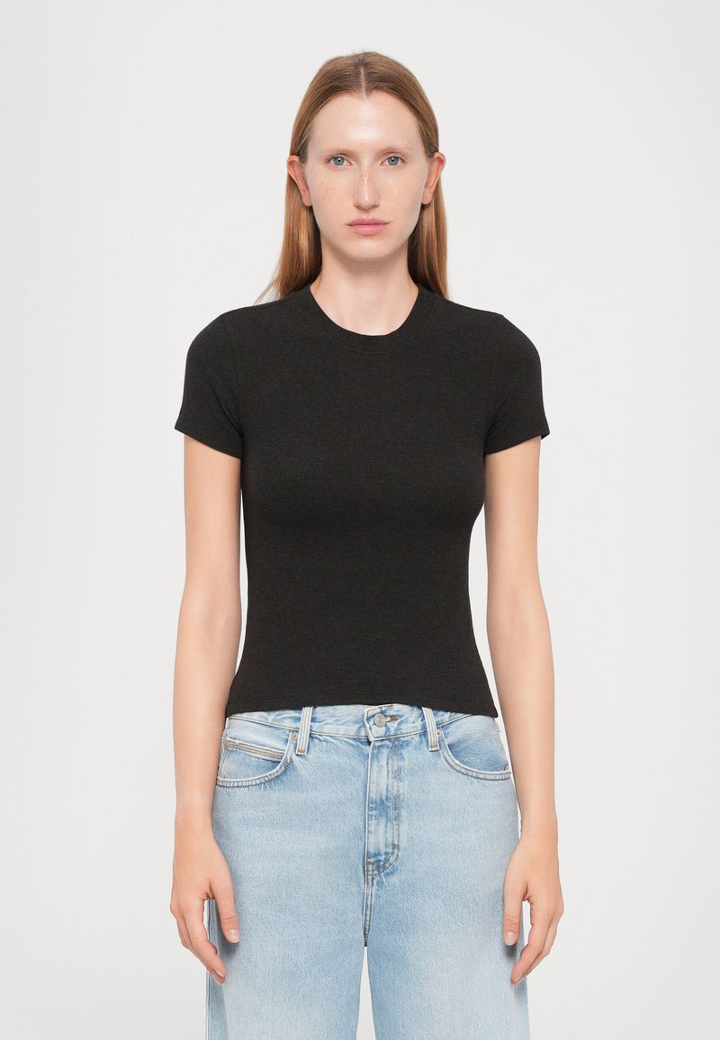 Young woman with straight red hair wearing a fitted black short-sleeve top and light blue high-waisted jeans, standing against a white background.