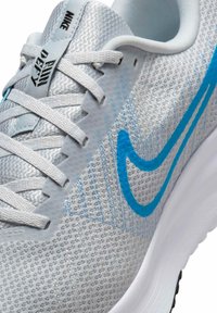 But sportowy Nike z siateczkową srebrną cholewką, niebieskim logo Swoosh, teksturowanymi detalami i białą podeszwą. Posiada płaskie sznurowadła i wyściełany kołnierz.