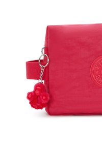 Borsa in tessuto rosso con superficie texturizzata, dotata di un logo circolare e di un charm decorativo in peluche rosa attaccato alla tracolla.