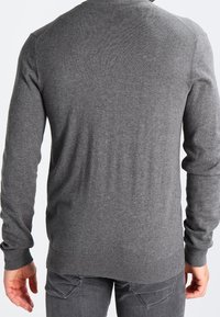Man som bär en långärmad grå tröja och mörkgrå jeans, sedd bakifrån mot en enkel vit bakgrund.
