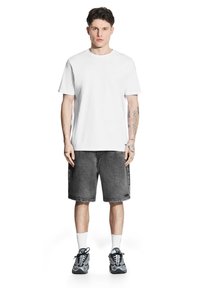 Biała bawełniana koszulka, czarne dżinsowe szorty, szare trampki, białe skarpetki do kostek. Model ma tatuaże na ramionach, stoi w neutralnej pozycji.