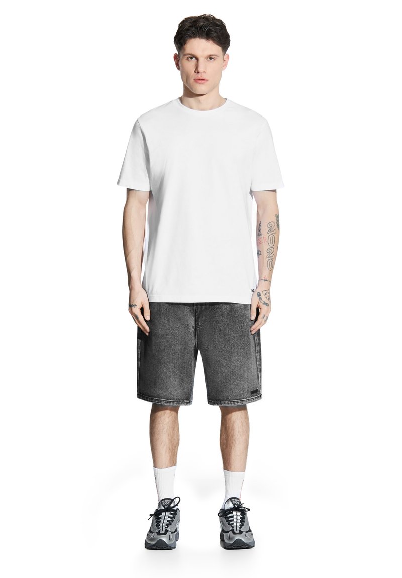 Biała bawełniana koszulka, czarne dżinsowe szorty, szare trampki, białe skarpetki do kostek. Model ma tatuaże na ramionach, stoi w neutralnej pozycji.