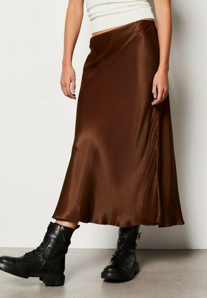 Osoba ubrana w brązową satynową spódnicę midi w połączeniu z czarnymi glanami na sznurowadła, stojąca na tle gładkiej, kremowej ściany.