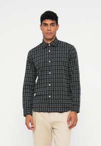 Man in een zwart-wit geblokt overhemd met beige broek, staand tegen een effen witte achtergrond.