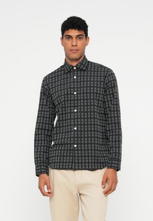 Hombre vistiendo una camisa de cuadros en blanco y negro con pantalones beiges, de pie frente a un fondo blanco liso.