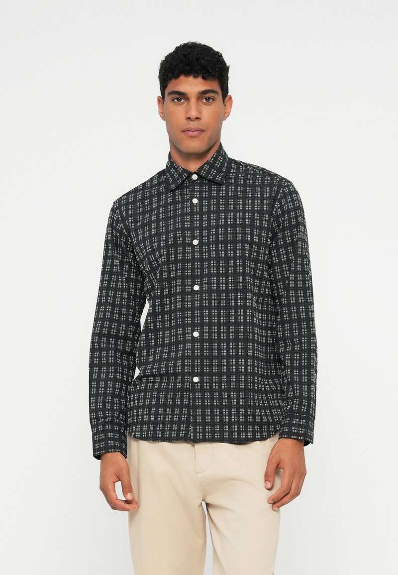 Man in een zwart-wit geblokt overhemd met beige broek, staand tegen een effen witte achtergrond.