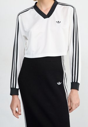Osoba nosząca biały krótki top z długim rękawem i czarną spódnicę, oba z czarnym wzorem trzech pasków adidas i logo.