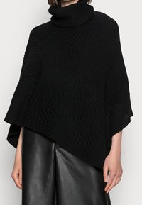 Femme portant un poncho à col roulé en maille noire et un pantalon en cuir noir, visible des épaules jusqu'au milieu des cuisses sur un fond uni.