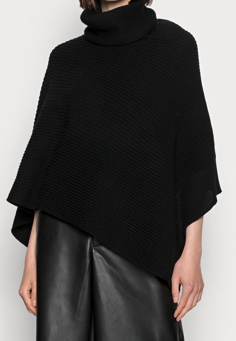 Femme portant un poncho à col roulé en maille noire et un pantalon en cuir noir, visible des épaules jusqu'au milieu des cuisses sur un fond uni.