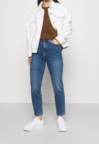 Chaqueta de mezclilla blanca sobre un suéter marrón, combinada con jeans azul de cintura alta y zapatillas blancas. Diseño simple, corte clásico, look casual.