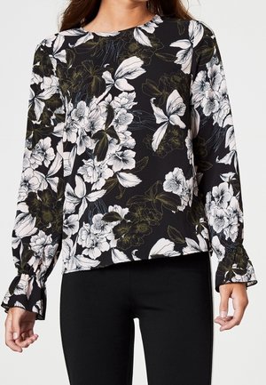 Zwarte blouse met lange mouwen en witte en groene bloemenprint, met gerimpelde manchetten, gedragen met zwarte getailleerde broek.