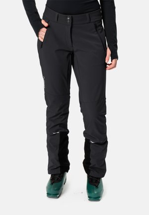 Pantaloni da sci neri con un design aderente, dotati di tasche laterali, una tasca con zip e polsini rinforzati sopra gli stivali da sci verdi.