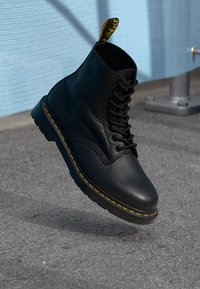 Zwarte leren veterschoen met gele stiksels en een gebrandmerkte pull-tab, zwevend boven het asfalt nabij een lichtblauwe muur.