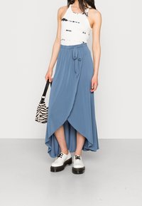 Blauwe wikkelrok met een high-low zoom, gecombineerd met een witte tanktop met een tie-dye patroon. Zwarte veterschoenen op een plateau en een gestreepte tas.
