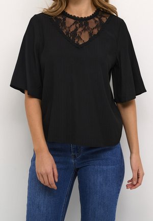 Femme portant un chemisier noir avec un col en V en dentelle et des manches courtes larges, associé à un jean denim bleu.