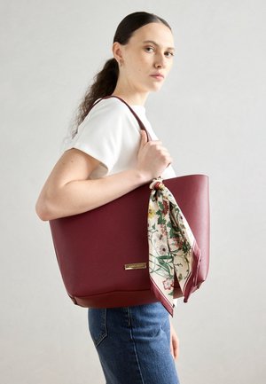 Vrouw met donker haar in een paardenstaart houdt een grote bordeauxrode boodschappentas vast met een bloemen sjaal vastgeknoopt aan het handvat, draagt een wit shirt en blauwe spijkerbroek.