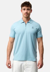 Lyseblå poloshirt med korte ærmer. Har en krave og et lille sort logo på brystet. Stoffet ser glat og let tekstureret ud.