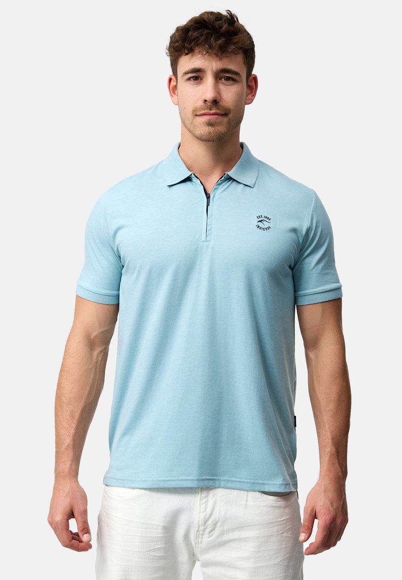 Lyseblå poloshirt med korte ærmer. Har en krave og et lille sort logo på brystet. Stoffet ser glat og let tekstureret ud.