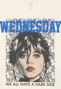 Krèmefarget t-skjorte med et grafisk trykk av en jente med mørkt hår og sterke røde lepper, teksten "WEDNESDAY" i blått over, og et blonderaktig bakgrunn.