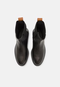 Melna ādas potīšu zābaku pāris ar noapaļotiem purngaliem, elastīgām sānu daļām un brūniem aizsējošajiem cilpiņām, skatīts no augšas uz baltā fona.