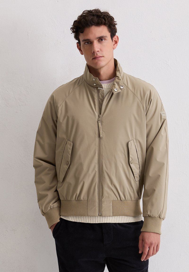 Beige bomberjack met een opstaande kraag, rits aan de voorkant en steekzakken. Heeft geribbelde manchetten en zoom, een gladde textuur en versterkte naden.