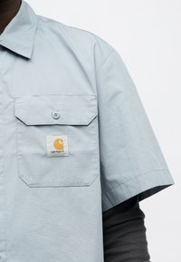 Φωτεινό γαλάζιο κοντομάνικο πουκάμισο με κουμπωτή τσέπη στο στήθος που φέρει μπάλωμα με το λογότυπο Carhartt, φορεμένο πάνω από μαύρο μακρυμάνικο τοπ.