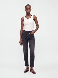 Femme portant un haut blanc sans manches avec un liseré noir, un jean noir taille haute et des chaussures Mary Jane rouge foncé, debout avec une main dans la poche.