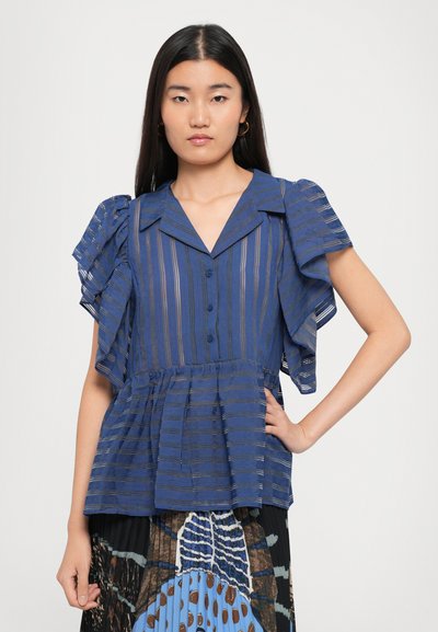 Woman with long hair wearing a blue striped blouse with ruffled sleeves and a patterned pleated skirt, posing with one hand on hip.