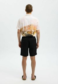 Košile s krátkým rukávem a knoflíky s akvarelovým designem městské krajiny v jemných pastelových barvách, doplněná černými šortkami a sandály.