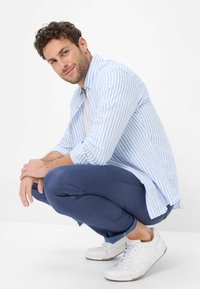 Camicia in cotone a righe azzurre e bianche sovrapposta a una t-shirt bianca, abbinata a pantaloni blu navy e sneakers bianche. L'uomo è accucciato con le mani giunte.