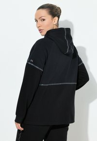 Sudadera con capucha negra con un acabado texturizado y acentos reflectantes en el pecho y los hombros. Mangas largas y un ajuste relajado.