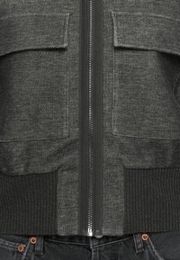 Vue de face d'une veste en maille gris foncé zippée avec deux poches à rabat et un ourlet côtelé sur un jean en denim noir.