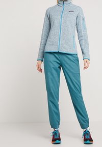 Vrouw in een lichtblauwe fleecejas met rits, teal joggerbroek en meerkleurige sportschoenen staat tegen een effen achtergrond.