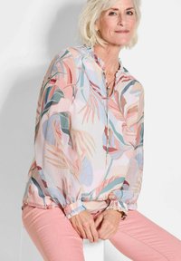 Langarmhemd aus leichtem, pastellfarbenem Stoff mit einem botanischen Druck von Blättern, gerafftem Ausschnitt und elastischen Bündchen. Kombiniert mit rosa Hosen.