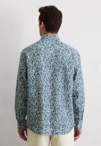 Chemise à manches longues avec un motif de feuilles bleu et crème, fabriquée en tissu léger, avec un col boutonné et un ourlet arrondi.